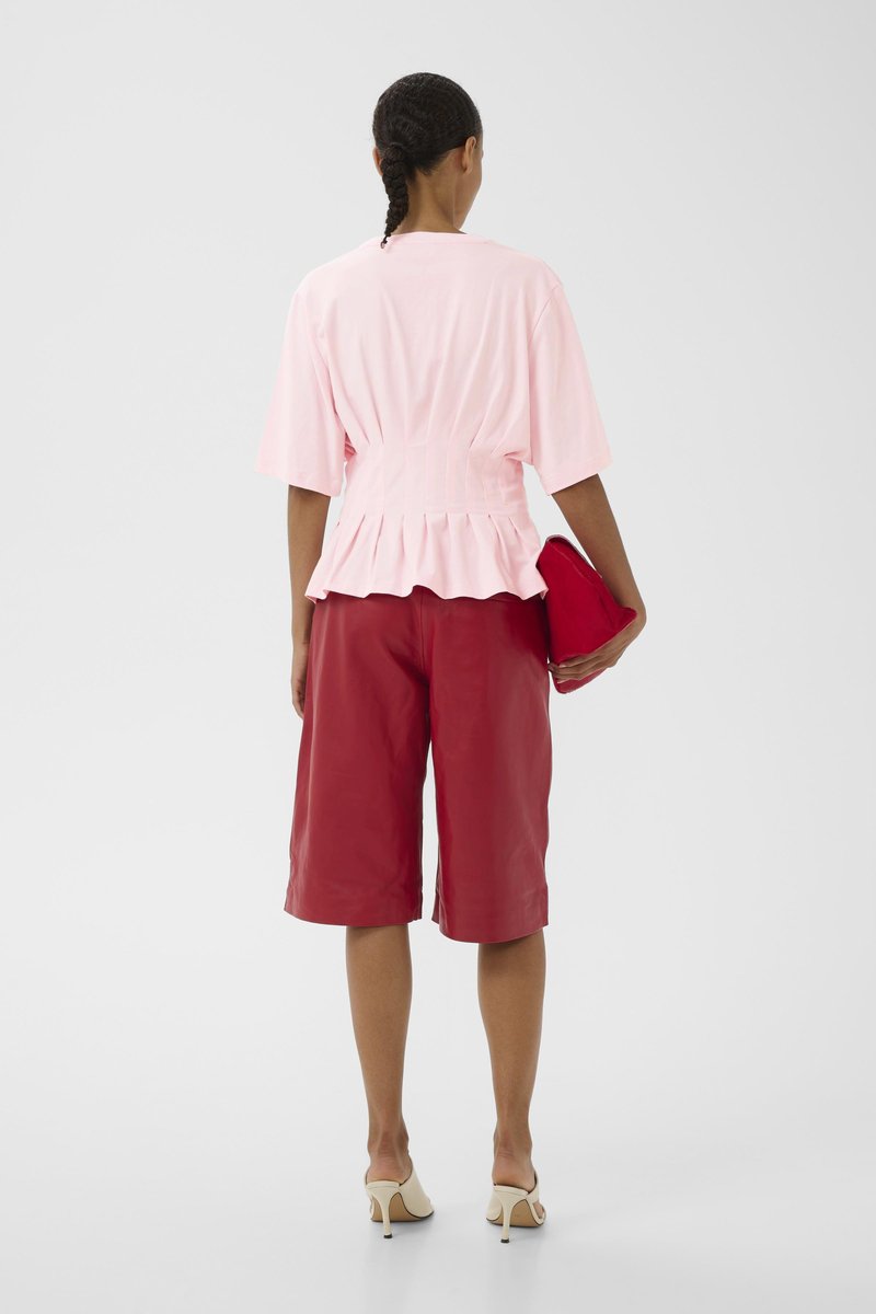 Kvinde med flettet hår iført en pink kortærmet top, røde shorts til knæet, beige hæle, og holder en rød clutch, vendt væk.