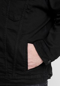 Black denim jacket with a chest pocket, featuring a textured fabric, button closure, and a hand resting in a side pocket.