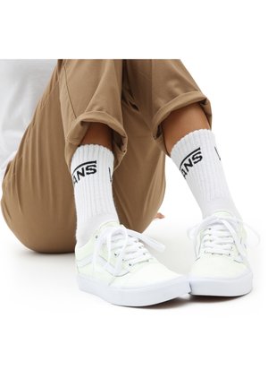 Zapatillas de lona blancas con un sutil brillo, con cordones, combinadas con calcetines blancos de canalé que tienen un logo negro de "VANS", llevadas con pantalones marrón claro.