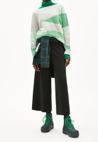 Pull oversize à rayures vertes et grises, pantalon large noir, et bottes bleu sarcelle avec des lacets verts ; chemise en flanelle superposée attachée à la taille.