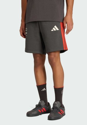 Mann trägt dunkelgraue Adidas-Shorts mit roten Streifen, passende Socken und schwarze Adidas-Sneaker mit roten Details, steht vor einem einfarbigen Hintergrund.