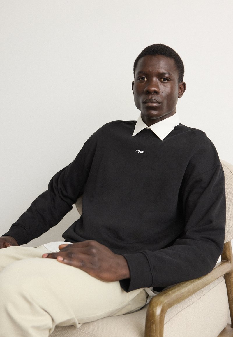 Felpa nera con logo "HUGO", sovrapposta a una camicia bianca con colletto. Modello seduto su una sedia chiara, indossando pantaloni beige.