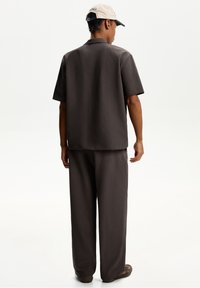 Brun texturerad kortärmad skjorta och matchande lösa byxor, tillsammans med en beige keps, sedd bakifrån, med en avslappnad passform.