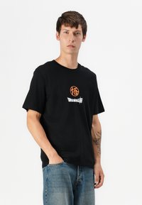 Fekete pamut póló rövid ujjakkal, narancssárga logóval és fehér "DRAGONBALL" felirattal a mellkason. Lazán illeszkedő, puha tapintású.