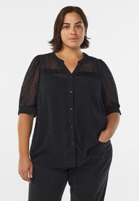 Blouse noire transparente avec des manches courtes bouffantes, motif texturé à pois, fermeture à boutons sur le devant et décolleté en V. Tissu doux avec une coupe ample.
