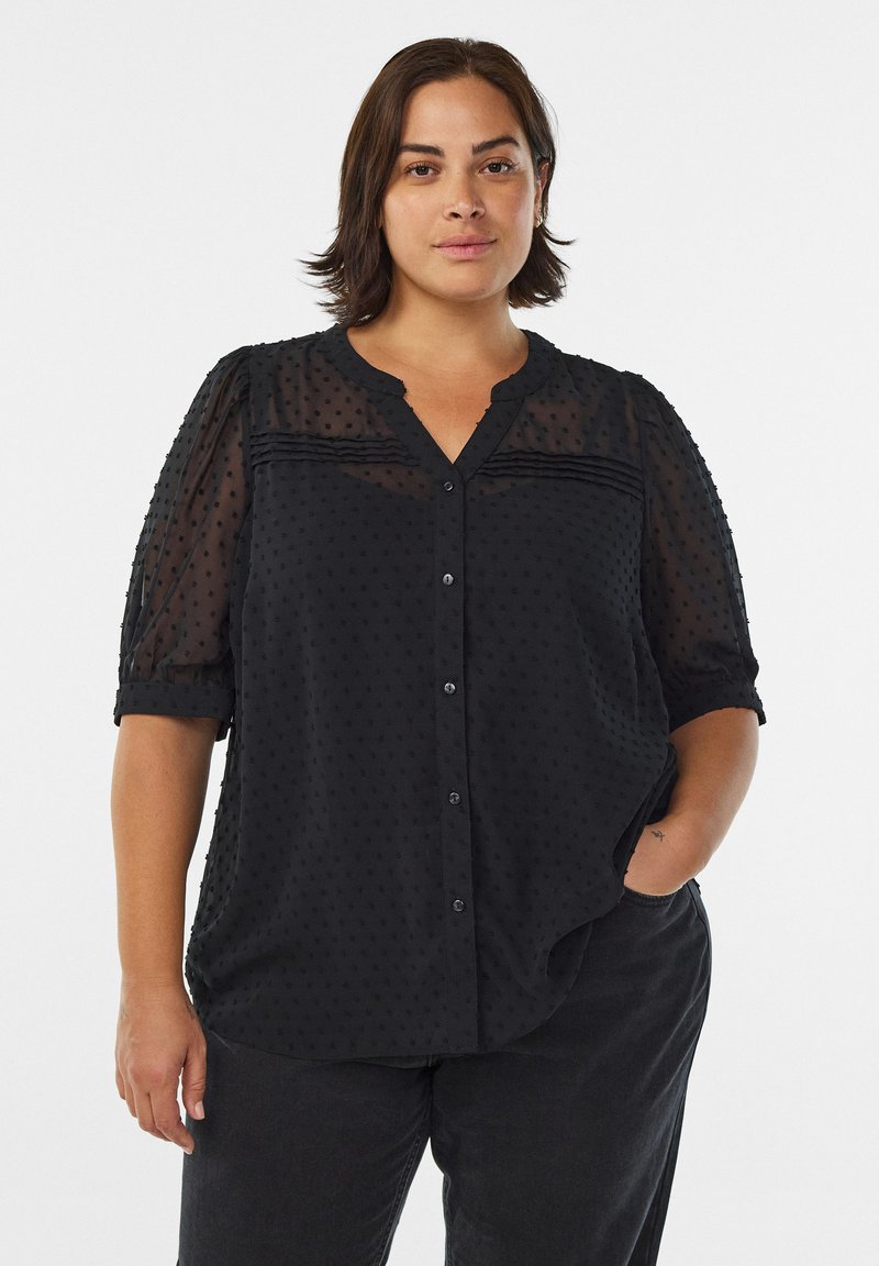 Blouse noire transparente avec des manches courtes bouffantes, motif texturé à pois, fermeture à boutons sur le devant et décolleté en V. Tissu doux avec une coupe ample.