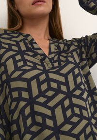 Langärmliges Oberteil aus glattem, olivgrünem Stoff mit einem geometrischen, dunkelblauen Muster, V-Ausschnitt und lockerer Passform.