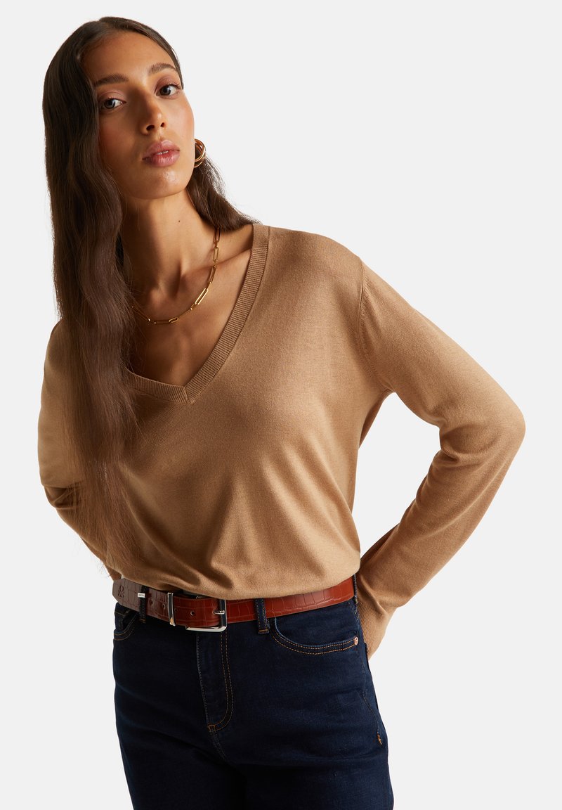 Beżowy sweter z dekoltem w kształcie litery V wykonany z lekkiego materiału, z długimi rękawami i luźnym krojem, połączony z ciemnym dżinsem i brązowym paskiem z tłoczeniem w kształcie krokodyla.