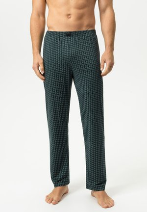 Mann trägt dunkle gemusterte lange Lounge-Hose, steht barfuß vor einem schlichten hellen Hintergrund.
