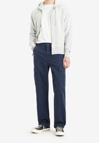 Grijze hoodie met rits en trekkoorden, wit T-shirt, marineblauwe cargobroek met zijzakken en zwarte sneakers met witte zolen.