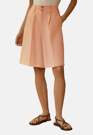 Kobieta ubrana w brzoskwiniowe, wysokowalne szorty za kolano z plisami i podwójnym zapięciem na guziki, zestawione z brązowymi sandałami i białym topem.