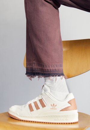 Bílá teniska Adidas s růžovými akcenty, nošená s bílými ponožkami a fialovými rozdrcenými kalhotami, odpočívající na světlé dřevěné židli.