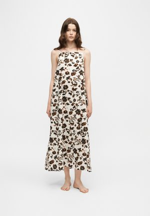 Jeune femme debout pieds nus portant une robe longue sans manches blanche avec un imprimé floral et animalier noir et marron sur fond uni.