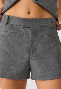 Pantaloni corti grigi su misura realizzati in un tessuto testurizzato. Le caratteristiche includono una vita piatta, due tasche frontali e un taglio dritto.