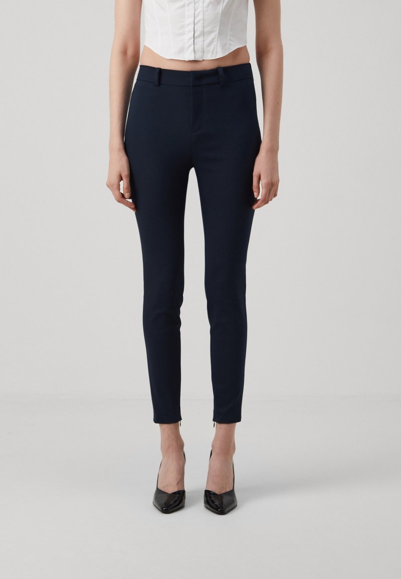 Femme portant un pantalon slim bleu marine aux chevilles, un top blanc court sans manches et des escarpins noirs pointus, debout devant un fond gris uni.