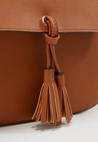 Hnědá kožená kabelka se střapcovým detailem a dvojitým třásním visícím z prošívaného popruhu provlečeného kovovou oční skobou na hladkém koženém povrchu.