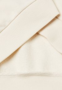 Tissu couleur crème avec une texture douce et des poignets côtelés. Présente une transition fluide entre les matériaux et un ourlet cousu.