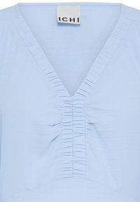 Blouse bleu clair avec encolure en V présentant des détails froncés et fripés le long de l'encolure et de la couture centrale avant.