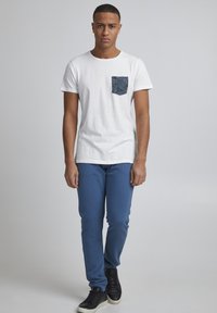 Camiseta de algodón blanca con un bolsillo estampado en azul, combinada con pantalones elásticos azules y zapatillas negras, de pie contra un fondo neutro.