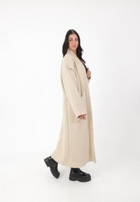 Cappotto lungo beige con maniche ampia, realizzato in materiale morbido. Ha tasche laterali profonde e una vestibilità comoda. Stivali neri con suola spessa.