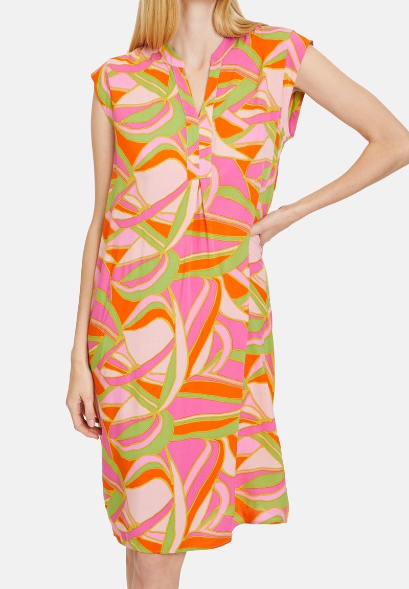 Femme portant une robe sans manches, longueur genou, avec un motif abstrait de feuilles orange vif, rose et vert, debout une main sur la hanche.
