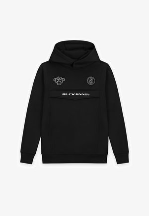 Schwarzer Hoodie mit Logodetails, kombiniert mit hellblauen, zerrissenen Jeans mit Farbklecksen und weißen Sneakern. Ein schwarzes Beanie rundet den Look ab.