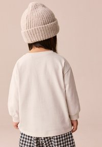 White, long-sleeve sweatshirt with ribbed cuffs, paired with checkered pants. A light gray knitted beanie covers the hair at the back.