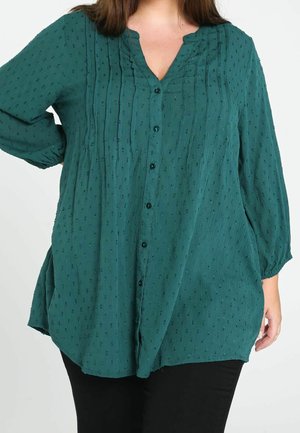 Donna che indossa una camicetta larga color teal con puntini tridimensionali, maniche lunghe, chiusura con bottoni sul davanti e piccole pieghe verticali vicino al collo.