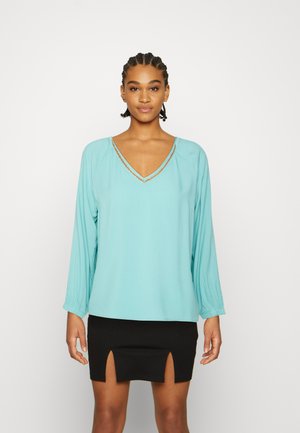 Blusa de mangas longas em azul-turquesa claro com decote em V e acabamento decorativo. Usada com uma saia mini preta com fendas frontais. Textura suave.