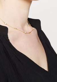 Collier chaîne en or avec des perles blanches espacées uniformément, porté autour du cou d'une personne vêtue d'un haut noir texturé à col en V.