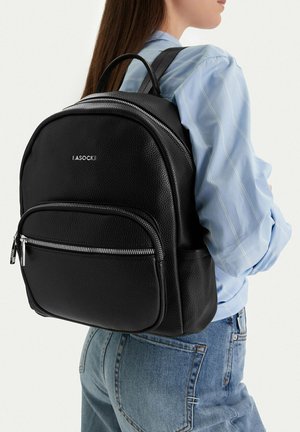 Mochila de cuero texturizado negro con bolsillo frontal con cremallera, usada por una persona con camisa de rayas azules y pantalones vaqueros azules, vista de perfil.