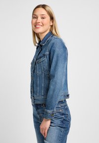 Joven sonriente, vestida con chaqueta vaquera azul y jeans, de pie de lado contra un fondo claro y liso.