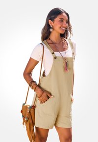 Khaki overall shorts van een lichte stof met twee vo pockets, gecombineerd met een wit t-shirt en een bruin schoudertasje.