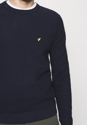 Man draagt een marineblauwe, getextureerde trui met lange mouwen en een klein geel vogel-logo op de linkerborst over een wit shirt met ronde hals.