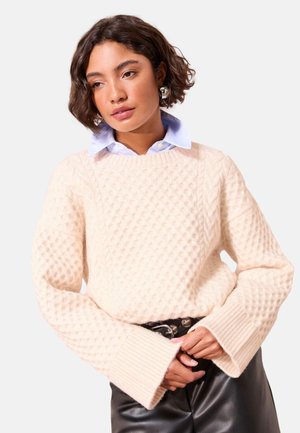 Mujer con pelo corto y rizado lleva un suéter texturizado color crema sobre una camisa azul claro con cuello, con pantalones de cuero negro y cinturón.