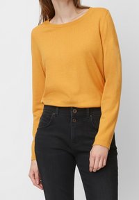 Pull jaune à manches longues avec un col rond, texture douce et coupe décontractée, associé à un jean noir avec une fermeture à deux boutons à l'avant.