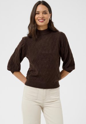 Femme souriante, portant un pull en maille marron texturé aux manches trois-quarts et un pantalon beige clair, debout devant un fond uni.