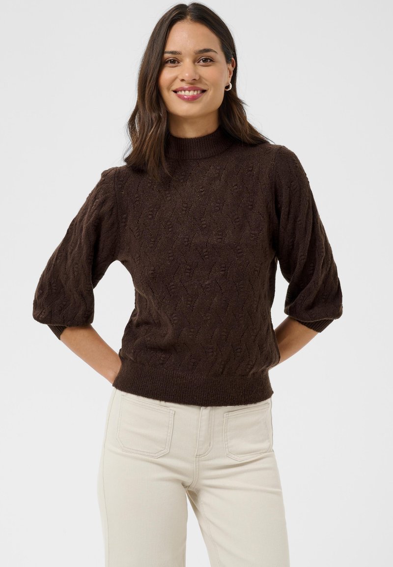 Femme souriante, portant un pull en maille marron texturé aux manches trois-quarts et un pantalon beige clair, debout devant un fond uni.