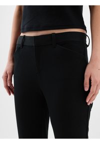 Personne portant un pantalon noir moulant et un top court noir, montrant le ventre, les mains reposant sur les côtés sur fond blanc.