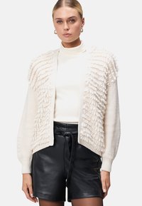 Crème cardigan met textuur, donzige details op de schouders, gebreide stof, open voorkant en lange mouwen, gecombineerd met hoge, zwarte shorts.