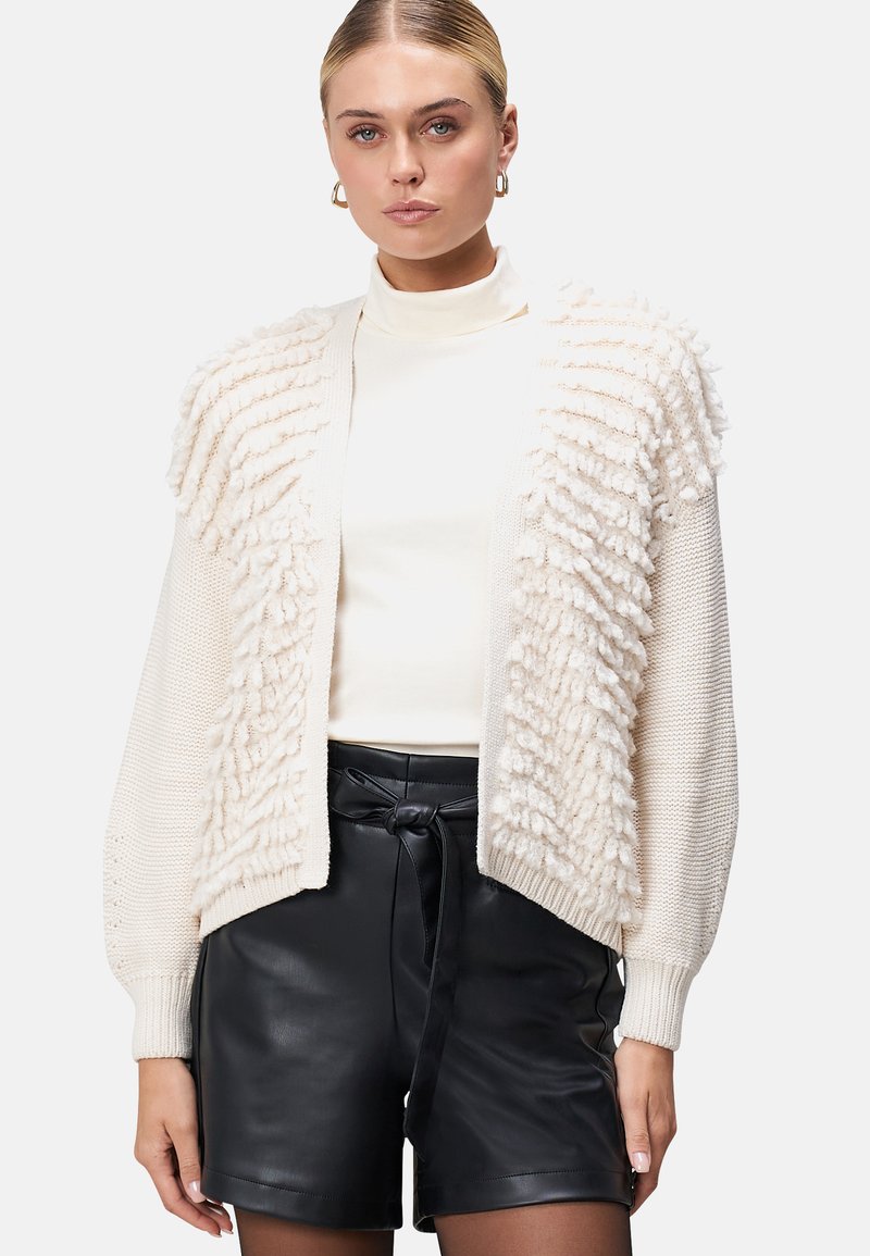 Crème cardigan met textuur, donzige details op de schouders, gebreide stof, open voorkant en lange mouwen, gecombineerd met hoge, zwarte shorts.