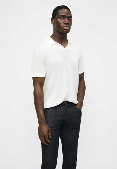 Άνδρας που στέκεται με το ένα χέρι στην τσέπη, φοράει κοντομάνικο λευκό πόλο πουκάμισο και σκούρο τζιν, μπροστά σε απλό λευκό φόντο.