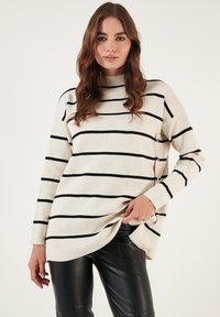 Maglione a righe beige con colletto alto, maniche lunghe e righe orizzontali nere. Indossato sopra pantaloni in pelle sintetica nera.
