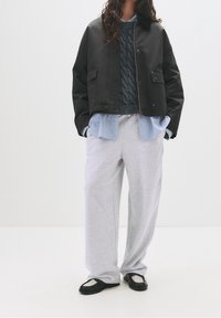 Veste noire raccourcie, pull gris en tricot torsadé, chemise rayée bleu clair, pantalon de survêtement gris ample, et chaussures plates noires avec des accents en maille.