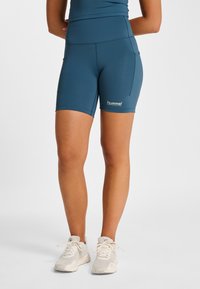 Teal atletiska shorts i stretchigt tyg, med hög midja, sidofickor och en liten vit logotyp på benet.