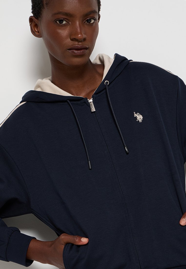 Vrouw in een marineblauwe hoodie met rits, voorzien van een beige capuchonvoering en een klein wit paardenlogo op de borst, met handen in de zakken tegen een vlakke achtergrond.
