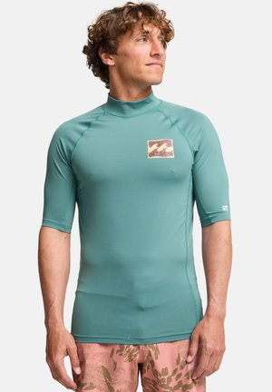 Blaugrünes Rashguard mit kurzen Ärmeln, hohem Kragen und einer strukturierten Markenlogo-Applikation auf der Brust. Dazu gemusterte rosa Badeshorts.