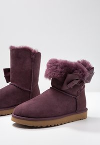 Bottes d'hiver en suède couleur prune, doublure en fourrure duveteuse, semelles en caoutchouc beiges basses, et accents de nœuds décoratifs sur les côtés arrière.