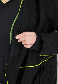 Ruka držící černý zip bundy poblíž hrudníku, oblečená v černém tričku s neonově zeleným lemováním a odpovídajícím neonově zeleným průvlakem na bundě.