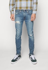 Vaqueros de mezclilla azul claro desgastados con corte ajustado, con roturas en las rodillas, textura desgastada y complementados con zapatillas verde-blancas.